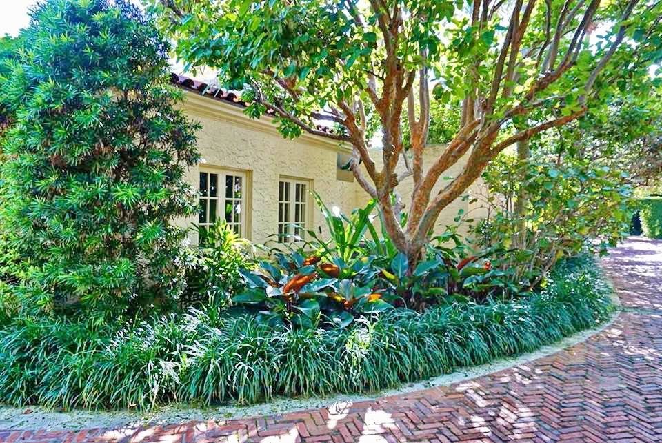 Brick walkway curving past layered tropical planting and mature trees beside a residence